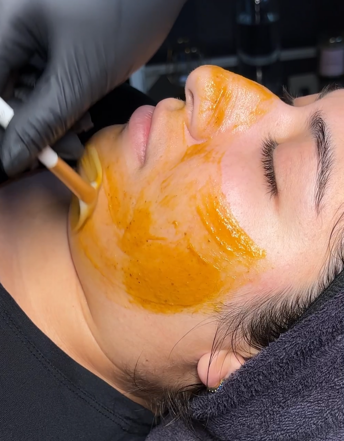 A gloved hand brushes a warm amber mask along a client's jawline during a facial at Lena Rose