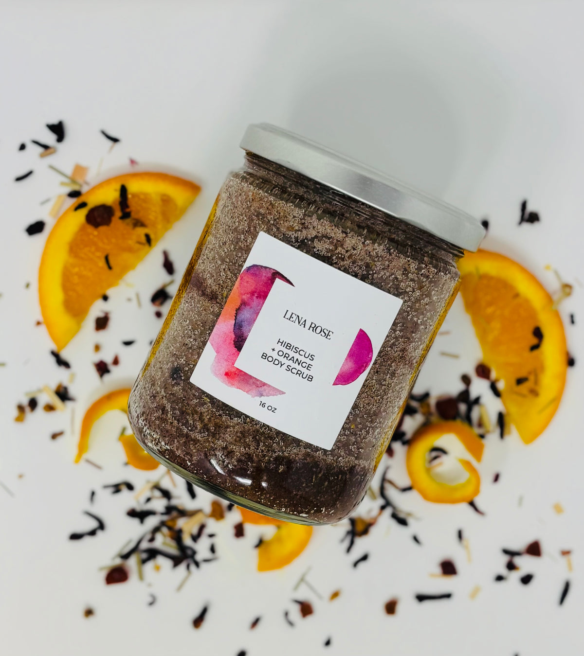 A jar of Lena Rose hibiscus and orange body scrub among dried orange slices and hibiscus petals