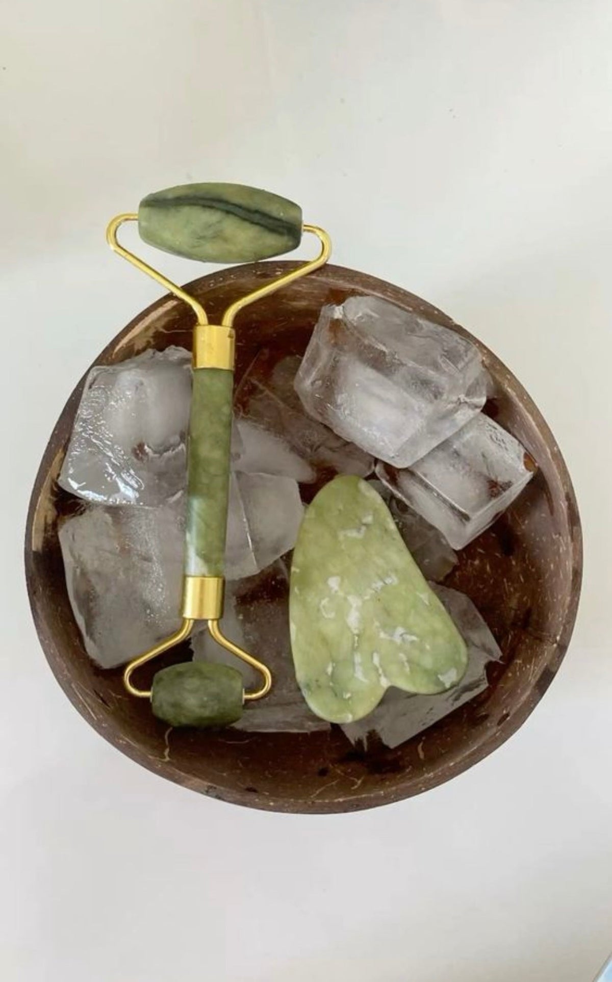 A jade face roller and gua sha stone resting in a wooden bowl of ice