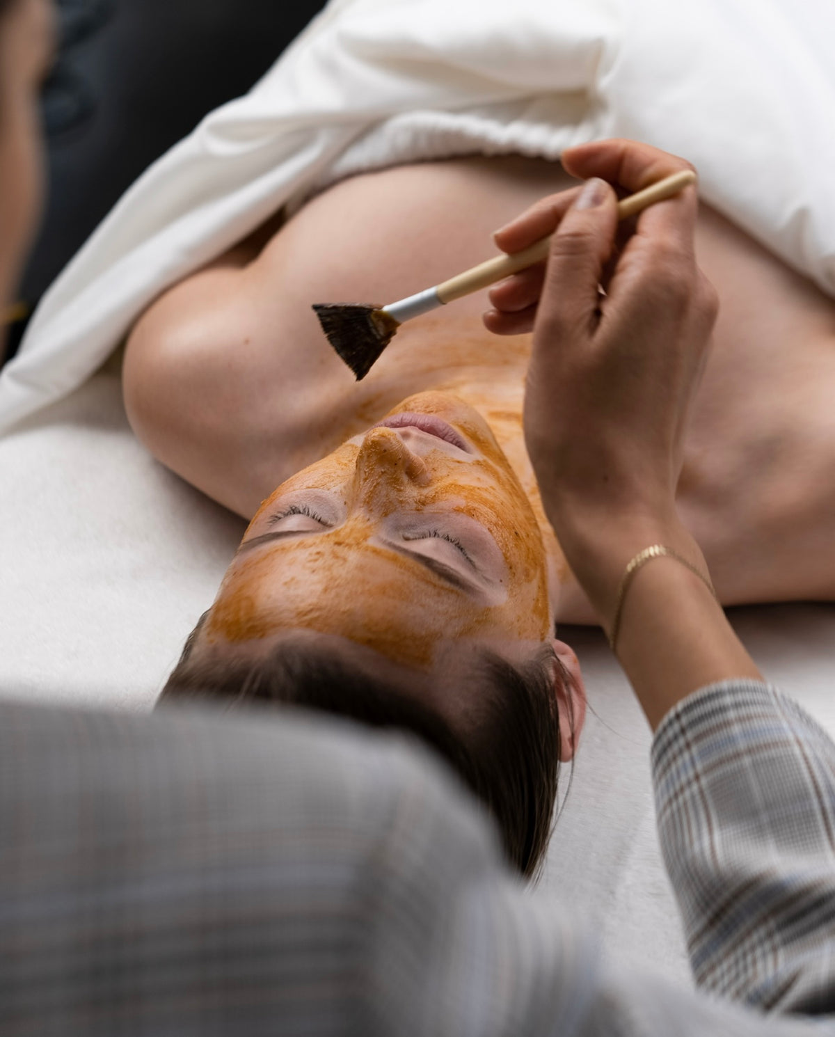 An esthetician brushes a warm amber mask across a client's face during a facial at Lena Rose.