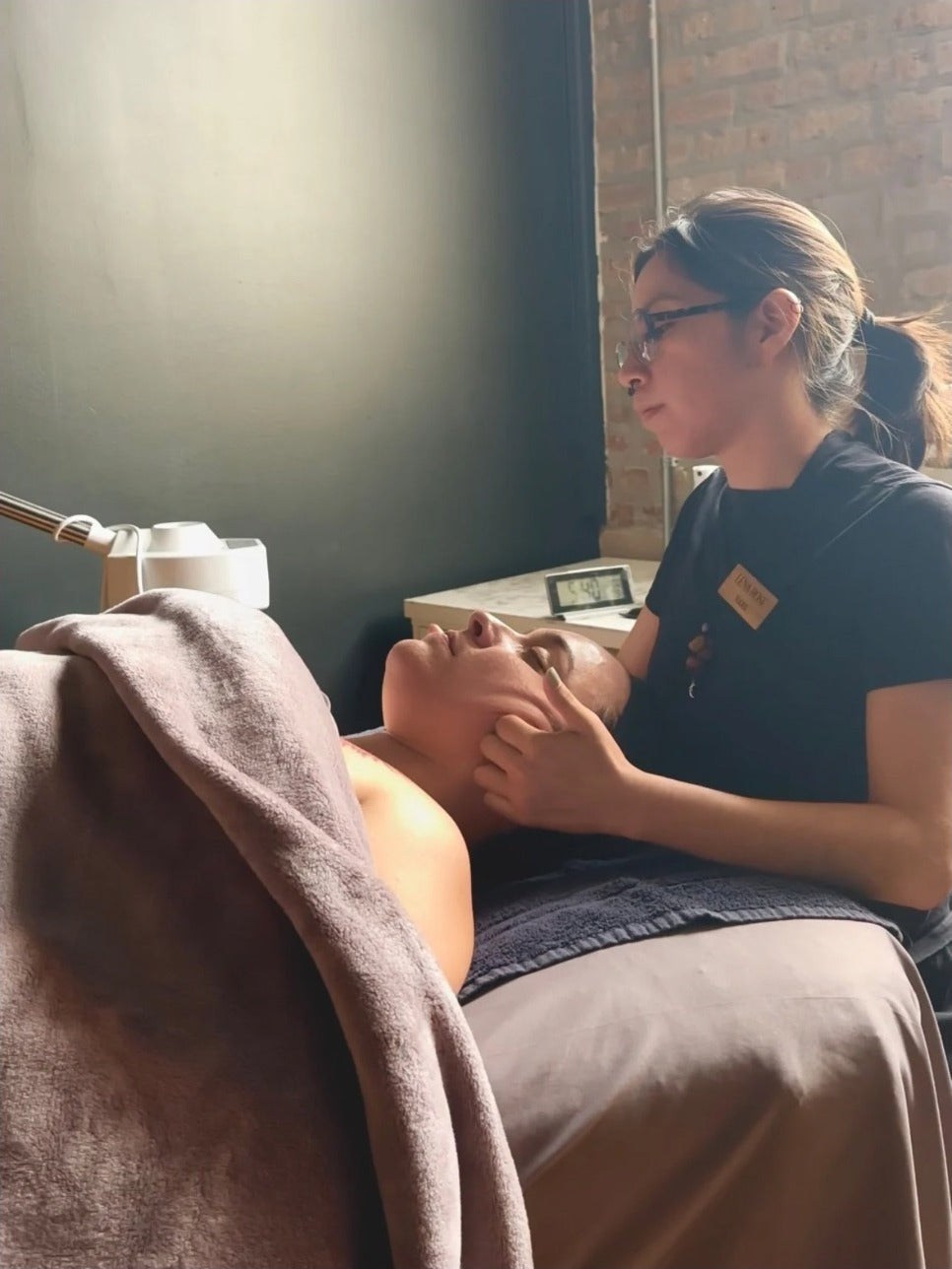 A Lena Rose esthetician supports a client's head during a facial in a brick-walled treatment room.